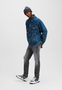 Chaqueta de mezclilla en azul oscuro con un corte oversized, dos bolsillos en el pecho y cierre de botones. Jeans grises desgastados y zapatillas con acentos azules completan el look.