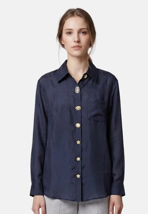 Femme portant un chemisier en soie bleu marine avec des boutons décoratifs uniques et une poche poitrine avant, associé à un pantalon gris clair.