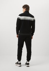 Svart velour treningsdress med høy krage og sølv horisontale striper på skuldrene, kombinert med matchende svarte joggebukser og sneakers.