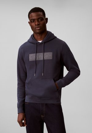 Młody mężczyzna ubrany w granatową bluzę z kapturem Calvin Klein z przednią kieszenią i ciemne dżinsy, stojący na tle jednolitego jasnego tła.