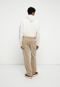 Pantaloni in corda beige con due tasche posteriori, abbinati a una felpa grigia chiara con testo nero sulla manica.