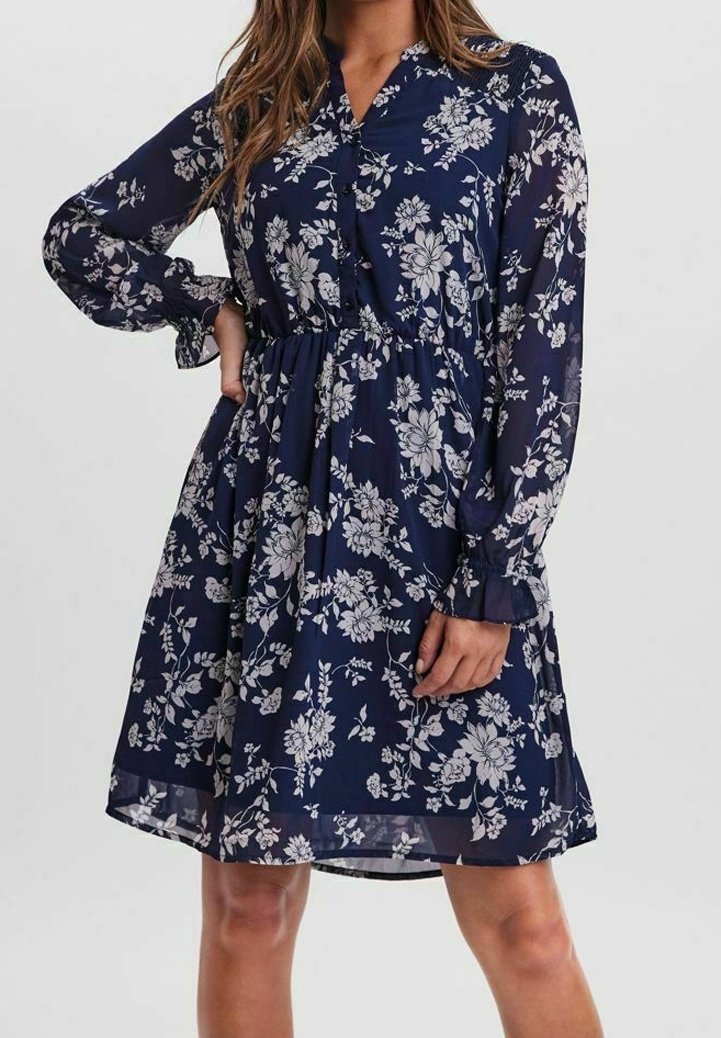 Robe bleu marine longueur genou avec motif floral blanc, manches longues transparentes, taille froncée et col en V portée par une personne.
