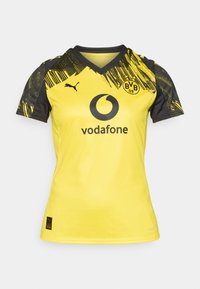 Maillot de sport jaune avec des accents noirs, design en V et motif frappant sur les manches. Comprend les logos d'une entreprise de téléphonie mobile et d'un club de football.