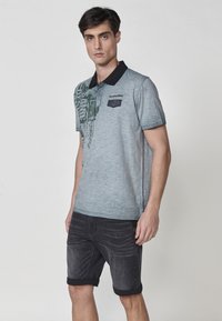 Sivo-heather polo majica s črnim ovratnikom, ki se ponaša z zelenim napisom in žepom na prsih z denim detajli, oblečena z temnimi kratkimi hlačami.