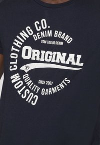 Camiseta de algodón azul marino con texto blanco desgastado: "Original," "Custom Clothing Co.," y "Quality Garments Since 2007." Diseño redondo.