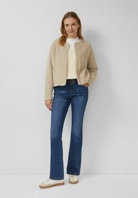 Beige gestrickte Strickjacke, weißer Pullover darunter, blaue ausgestellte Jeans und cremefarbene Sneaker mit braunen Sohlen. Schlichtes Design, strukturierter Strick.