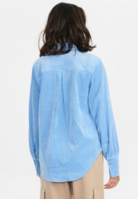 Camicia in tessuto morbido azzurro chiaro con polsini con bottoni, dettaglio pieghe sulla schiena e orlo curvo. Le maniche lunghe creano una silhouette rilassata.