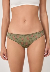 Woman wearing olive green bikini-style underwear with orange leopard and palm leaf print and a small front bow, shown from waist to thighs.
