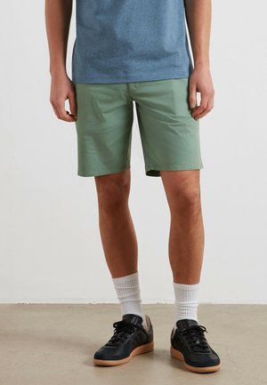 Uomo che indossa pantaloncini verdi fino al ginocchio, calzini bianchi alla caviglia, scarpe da ginnastica nere e una maglietta blu, in piedi su un pavimento chiaro contro una parete bianca.