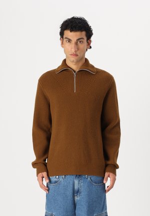 Brown ribbed knit sweater with a zippered collar, featuring vertical pattern detailing and long sleeves. Paired with blue denim shorts.