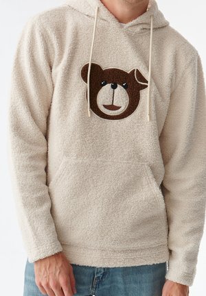 Beżowa bluza z kapturem wykonana z sherpy, z brązową twarzą misia na piersi, kapturem ze sznurkiem i kieszenią typu kangur z przodu, noszona z niebieskimi dżinsami.