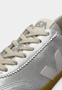 Baskets en cuir argenté avec lacets blancs et accents blancs, présentant une surface texturée et un logo latéral angulaire. Semelle extérieure en caoutchouc avec détails marron.