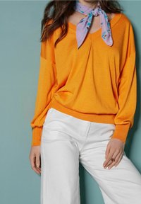 Orange stickad tröja med V-ringning och lös passform, i kombination med vita vida byxor. En ljuslila scarf är knutna runt halsen.