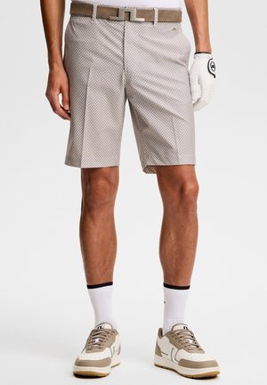 Mann trägt beige gemusterte Shorts, weißen Gürtel, weißen Golfhandschuh, weiße Socken und beige-weiße Turnschuhe.