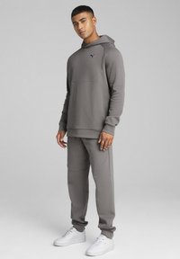 Grå sweatshirt med hætte og forlommer, parret med matchende bukser. Har en glat tekstur, sort logo og smalle manchetter. Hvide sneakers.