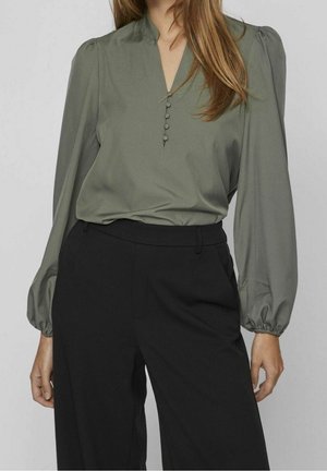 Femme portant un chemisier vert olive à manches longues avec des boutons recouverts de tissu et un pantalon noir taille haute à jambes larges.