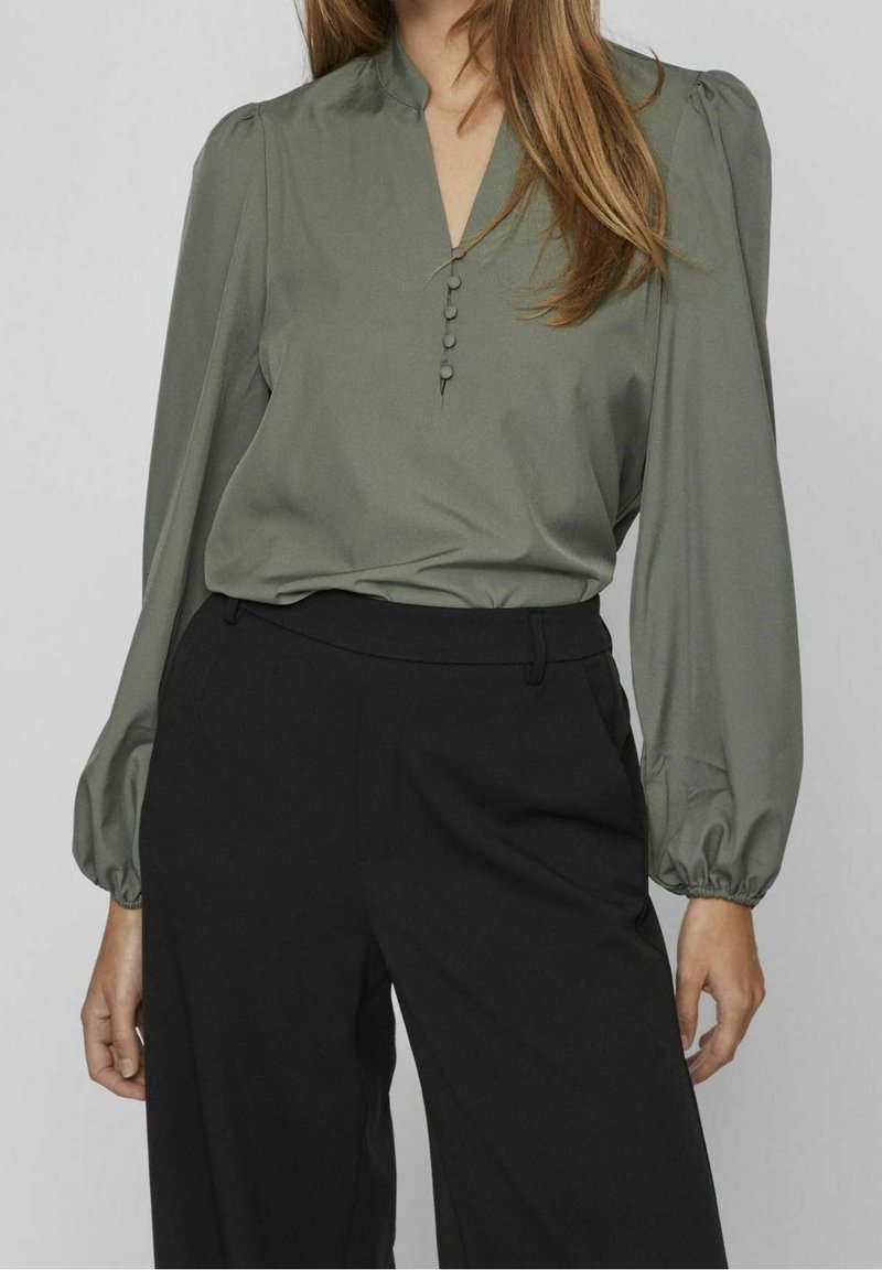 Femme portant un chemisier vert olive à manches longues avec des boutons recouverts de tissu et un pantalon noir taille haute à jambes larges.