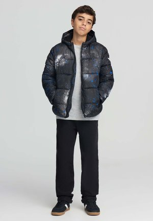 Ragazzo in piedi con le mani nelle tasche, indossa un giubbotto imbottito con motivo galattico nero e blu, una maglietta grigia, pantaloni neri e scarpe da ginnastica nere con strisce bianche.