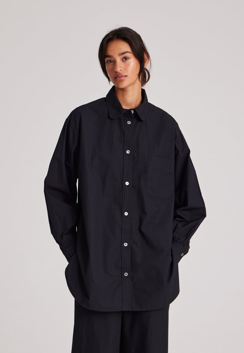 Camisa negra de botón con cuello, mangas largas y un bolsillo en el pecho. Hecha de una tela suave, con un corte holgado y botones plateados.