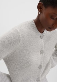 Vrouw in een lichtgrijze gebreide cardigan met grote knopen, staand met de handen op de heupen en het hoofd iets gebogen.