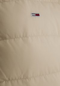 Beige pufferjack met een doorgestikt patroon, met een klein geborduurd logo in rood, wit en blauw op de bovenlinkerkant.