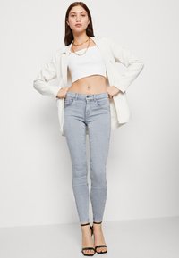 Jeans ajustados de color azul claro con un ligero estiramiento, que presentan una cintura de altura media, diseño de cinco bolsillos y dobladillo deshilachado. Combinados con una camiseta corta blanca y una chaqueta.