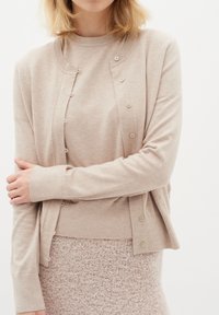 Beige Strickjacke mit rundem Halsausschnitt, Knopfleiste und gerippten Bündchen, getragen über einem strukturierten, hellrosa Pullover. Weiches Strickmaterial.