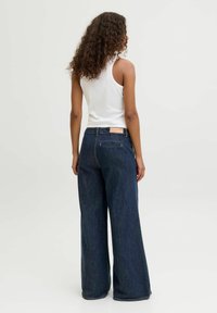 Young woman with curly hair wearing sleeveless white top and wide-leg dark blue denim jeans, standing facing away on plain background.