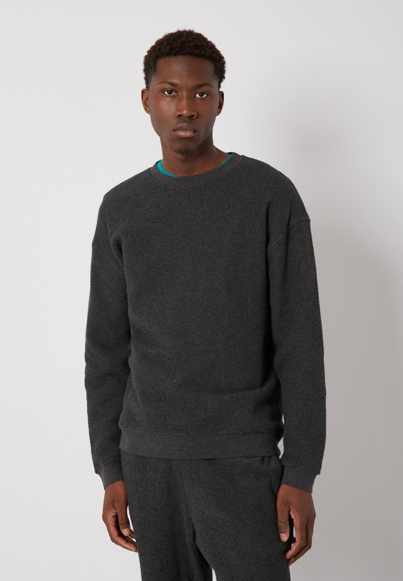 Mørkegrå crewneck sweatshirt med en blød, struktureret overflade. Har ribbet krave og manchetter. Båret over en turkise skjorte.