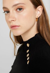 Schwarzer, gerippter Pullover mit hohem Kragen und fünf goldfarbenen Zierknöpfen an der Schulter. Modell mit braunen Haaren, im Profil gezeigt.