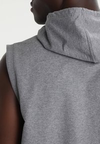 Primer plano de una persona con un chaleco gris sin mangas, mostrando la parte posterior y el perfil lateral del cuello y el hombro.