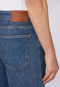 Mörkblå denimjeans med en lädertagg märkt "TRUSSARDI." Har en klassisk femfickedesign och kontrasterande sömmar.