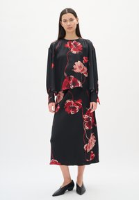 Donna che indossa un completo nero a due pezzi con motivi floreali rossi e rosa, maniche lunghe, una gonna midi e scarpe nere a punta.