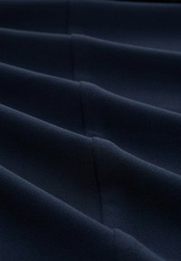 Dunkelblauer Stoff mit glatter Textur, der ausgeprägte, schräg verlaufende Falten und Nähte aufweist, die ein dynamisches, schichtartiges Erscheinungsbild erzeugen.