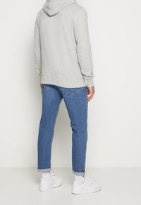 Personne debout, de dos, portant un sweat à capuche gris clair, un jean bleu retroussé, des chaussettes blanches et des baskets blanches, sur un fond uni.