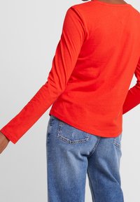 Rood katoenen shirt met lange mouwen, een ronde halslijn, een aansluitend silhouet en een gladde textuur. Draagt een lichtblauwe spijkerbroek met achterzakken.