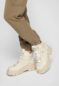 Baskets montantes beiges avec une finition en cuir texturé, semelle en caoutchouc épaisse et logos bien visibles. Portées avec un pantalon fuselé vert olive.