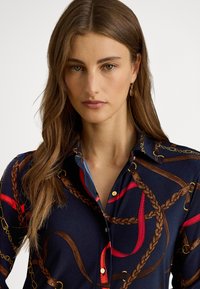 Jauna sieviete ar gariem brūniem matiem valkā tumši zilu blūzi ar brūnām pītām siksnām un sarkaniem akcentiem, zelta pogu detaļām un ovāliem auskariem.