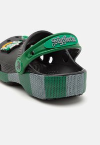 Černá bota ve stylu krokodýlí kůže se zeleným pásem Slytherinu a šedě pruhovanou podrážkou. Na svršku jsou dekorativní prvky a větrací otvory.