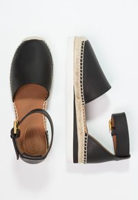 Espadrille de cuero negro con punta cerrada, correa en el tobillo y detalles de cuerda trenzada. Suela blanca, textura suave y forro interior marrón.