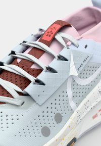 Chaussures de trail Nike avec une tige en mesh gris, des accents roses, des superpositions texturées et des lacets rouges, dotées de perforations pour la respirabilité.