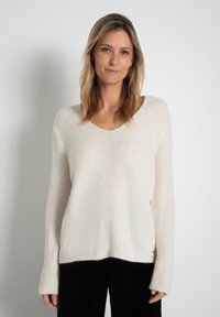 Cremefarbener Pullover mit V-Ausschnitt, strukturiertem, geripptem Strickmuster und langen, leicht ausladenden Ärmeln, kombiniert mit schwarzen Cordhosen.