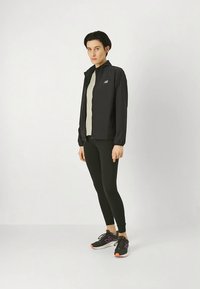 Zwarte lichtgewicht jas met een hoge kraag, gecombineerd met een nauwsluitend licht topje en zwarte leggings. Schoenen hebben paarse accenten en een gerubberde zool.