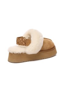 Suede platform slide shoe in tan, featuring a fluffy white shearling collar and a patterned tan strap with metal hardware. Textured rubber sole.