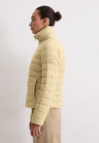 Lichtgele donzen jas met hoge kraag, doorgestikt patroon, zijzak en logo-accent op de mouw. Soepele, gewatteerde textuur.