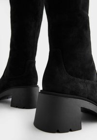 Zwarte suede kniehoge laarzen met een dikke, matte zwarte hak; gladde textuur met subtiele stiksels langs de naden.