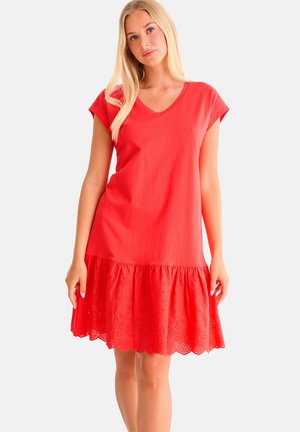 Jeune femme aux longs cheveux blonds portant une robe rouge à manches courtes avec un ourlet en dentelle ajourée festonnée, posant devant un fond blanc.