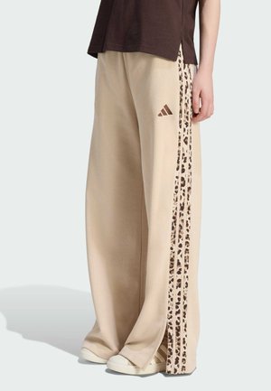 Beige broek met wijde pijpen, luipaardprint en drie strepen aan de zijkant, Adidas-logo op de dij, gedragen met witte sneakers en een donkerbruin topje.