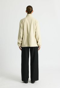 Beige Fleecejacke mit hohem Kragen und elastischen Bündchen, kombiniert mit lockeren schwarzen Hosen. Schlichtes Design, weicher Stoff, ohne Muster.