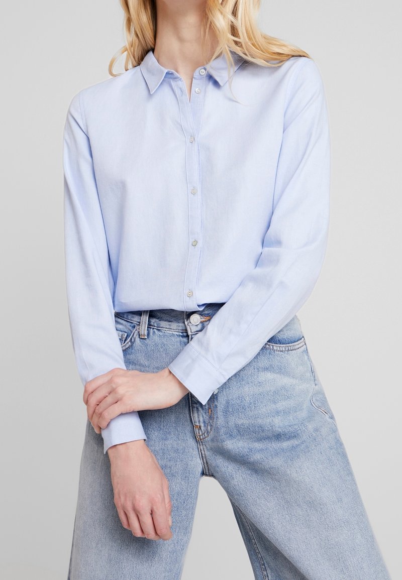 Femme portant une chemise bleu clair à manches longues boutonnée, rentrée dans un jean bleu clair taille haute, les mains doucement croisées.